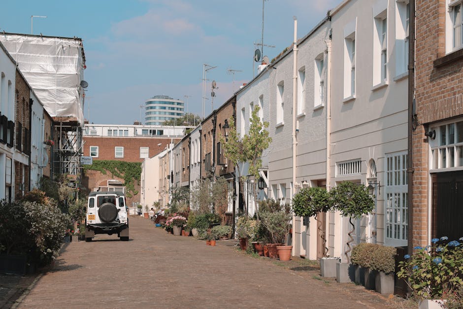 Image depicting a narrow residential street in Ealing with a white van parked on the cobblestone road, which is lined with various potted plants and small trees outside terraced houses. The houses hav