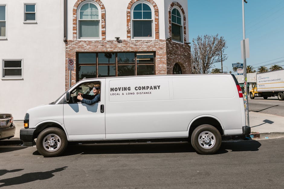 A man wearing a red cap, red T-shirt, and blue trousers stands outside a white panel van with its rear doors open, loading or unloading cardboard boxes and plastic crates filled with various items. Th